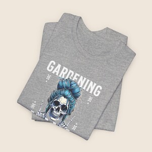 Pu&ograve; includere: Una maglietta grigia con la scritta "GARDENING" in lettere bianche. La maglietta presenta un teschio con i capelli blu raccolti in uno chignon, che fa un gesto con la mano rock and roll. Il design include piccoli motivi a diamante bianchi.