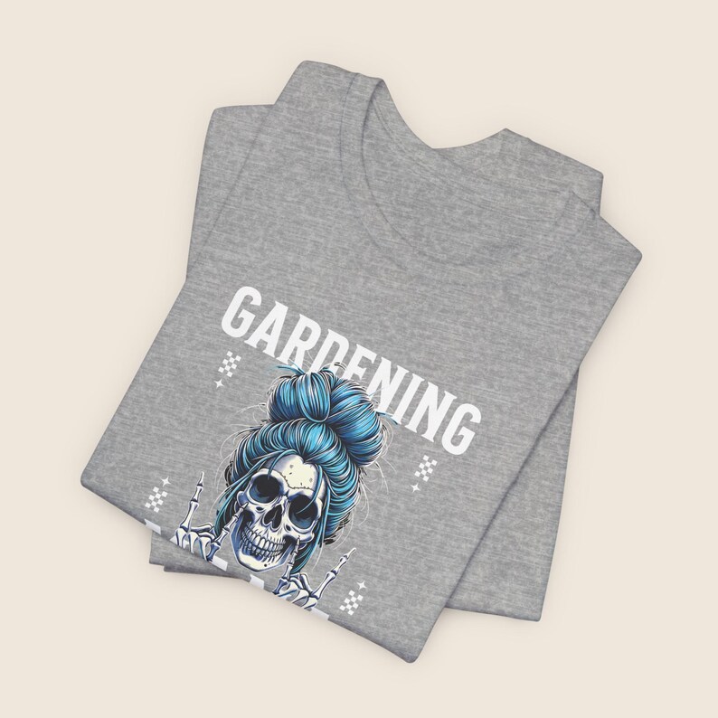 Pu&ograve; includere: Una maglietta grigia con la scritta "GARDENING" in lettere bianche. La maglietta presenta un teschio con i capelli blu raccolti in uno chignon, che fa un gesto con la mano rock and roll. Il design include piccoli motivi a diamante bianchi.