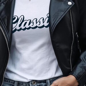 May include: A black leather jacket with silver zippers and snaps over a white t-shirt. The t-shirt has the word "Classi" in a blue script font. The model is wearing blue jeans and a layered necklace.