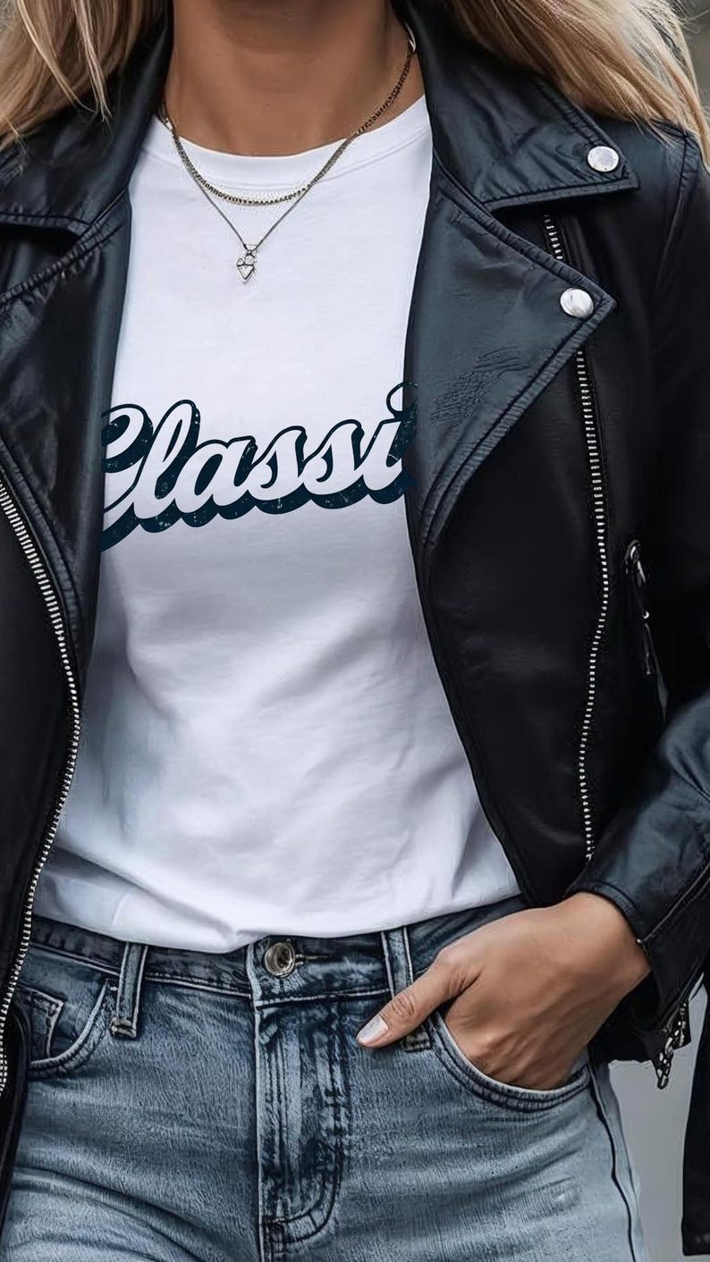 May include: A black leather jacket with silver zippers and snaps over a white t-shirt. The t-shirt has the word "Classi" in a blue script font. The model is wearing blue jeans and a layered necklace.
