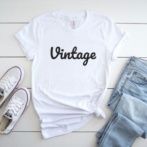 May include: White t-shirt with the word "Vintage" in black script. The shirt is paired with light blue jeans and white sneakers with red accents. The items are arranged on a white wooden surface.