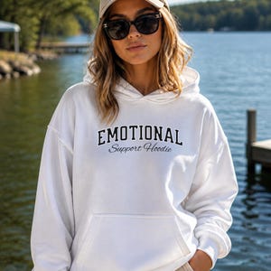 May include: A white hoodie with the text "EMOTIONAL Support Hoodie" in black. The hoodie has a front pocket and a drawstring hood. The image is taken outdoors with a lake in the background.