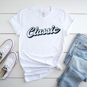 May include: White t-shirt with the word "Classic" in a navy blue script font. The shirt is paired with light blue jeans and white sneakers. The shirt is knotted at the waist.