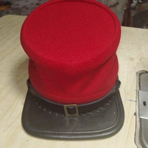 Könnte beinhalten: Ein roter Wollhut mit schwarzem Lederkrempe und einer goldfarbenen Schnalle. Der Hut hat eine abgerundete Oberseite und ein strukturiertes Design. Der Krempe ist genäht und hat einen Lederriemen.