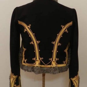May include: Black jacket with gold trim and gray fur collar and cuffs. The jacket has gold decorative trim along the front and back. The jacket is displayed on a wooden stand. The jacket is a vintage style.