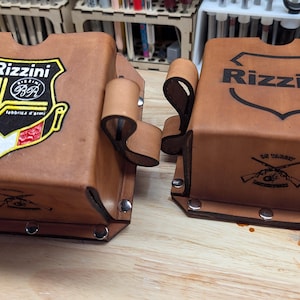 May include: Two brown leather tool holders with the name "Rizzini" and a logo. One has a colorful emblem, the other has a black logo. Both have loops and are riveted.