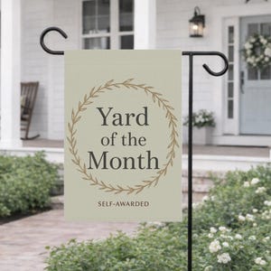 Può includere: Una bandiera da giardino beige con la scritta "Yard of the Month" all'interno di una ghirlanda marrone. Le parole "SELF-AWARDED" sono sotto il testo principale. La bandiera è appesa a un supporto di metallo nero davanti a una casa bianca.