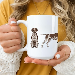 May include: A white ceramic mug featuring a detailed illustration of a German Shorthaired Pointer dog shown in two positions: sitting upright facing forward and standing in profile, both displaying a brown and white speckled coat pattern.