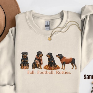 May include: A sand-colored crewneck sweatshirt featuring a graphic of four Rottweiler dogs in autumn-themed attire and football gear. Below the dogs, the text reads, Fall. Football. Rotties. The sweatshirt has a ribbed collar and long sleeves.