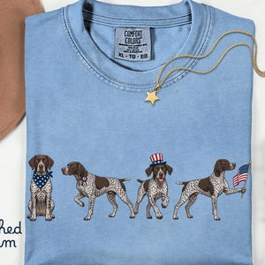 May include: Light blue t-shirt with a graphic of four dogs, each with patriotic accessories. The shirt has the words "Comfort Colors" and "XL - TG - EG" printed on the tag. A gold star pendant necklace is draped across the shirt.