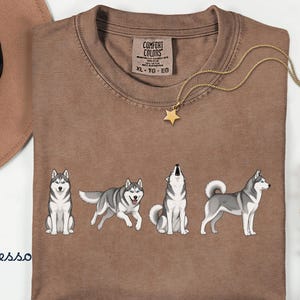 May include: Espresso-colored t-shirt featuring a graphic of four Siberian Huskies in different poses. The shirt has a crew neck and short sleeves. A gold necklace with a star pendant and a brown hat are also visible.