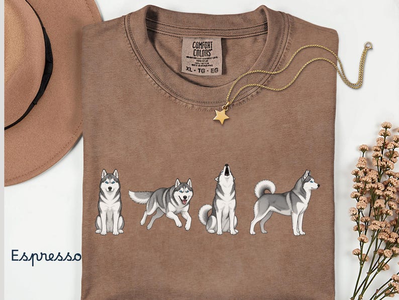May include: Espresso-colored t-shirt featuring a graphic of four Siberian Huskies in different poses. The shirt has a crew neck and short sleeves. A gold necklace with a star pendant and a brown hat are also visible.