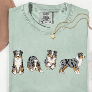 May include: A light green t-shirt featuring four embroidered Australian Shepherd dogs in various poses. A gold necklace with a star pendant and a brown hat are also visible. The shirt has a Comfort Colors tag.