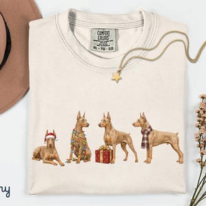 May include: An ivory t-shirt featuring a graphic of four Doberman dogs. One dog wears a Santa hat, one is wrapped in colorful Christmas lights, one stands by a red gift box, and one wears a plaid scarf. The shirt has a Comfort Colors label.