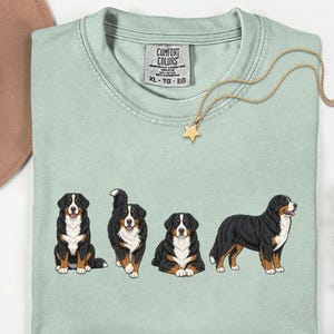 Op de afbeelding: Lichtgroen T-shirt met vier illustraties van Berner Sennenhonden. Het shirt heeft een Comfort Colors-label en een gouden ketting met een sterhanger. Een bruine hoed en bloemen zijn ook zichtbaar.