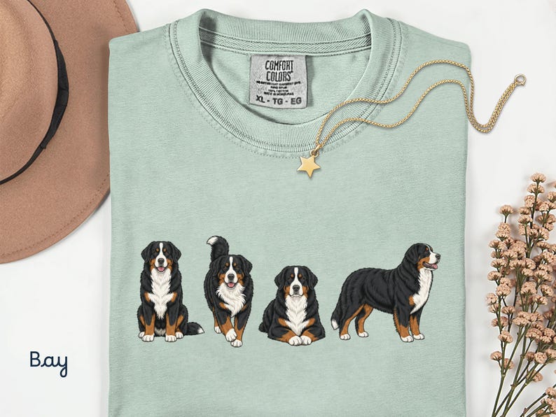 Op de afbeelding: Lichtgroen T-shirt met vier illustraties van Berner Sennenhonden. Het shirt heeft een Comfort Colors-label en een gouden ketting met een sterhanger. Een bruine hoed en bloemen zijn ook zichtbaar.