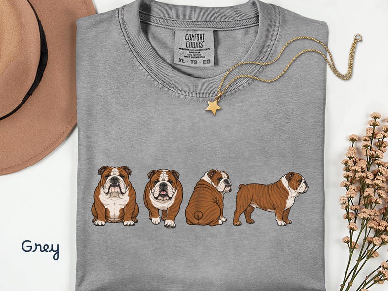 May include: A grey Comfort Colors t-shirt with a graphic of four brown and white bulldogs in various poses. A gold star pendant necklace is on the shirt. The word "Grey" is written in cursive in the bottom left corner.