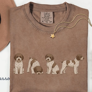 Può includere: Una t-shirt color espresso con una stampa grafica di quattro cuccioli di Lagotto Romagnolo in varie pose. La maglietta presenta uno scollo rotondo e un'etichetta del marchio Comfort Colors sul colletto interno. Il testo "Espresso" è stampato in blu a sinistra.