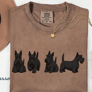 May include: Espresso-colored t-shirt featuring four black Scottish Terrier dog illustrations. The shirt has a crew neck and a tag that reads "Comfort Colors". A gold necklace with a star pendant is draped across the shirt.