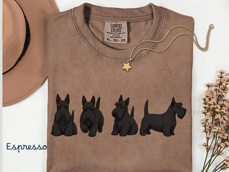 May include: Espresso-colored t-shirt featuring four black Scottish Terrier dog illustrations. The shirt has a crew neck and a tag that reads "Comfort Colors". A gold necklace with a star pendant is draped across the shirt.