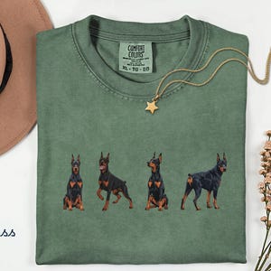 May include: A moss green t-shirt featuring a graphic print of four Doberman dogs in various poses. The shirt has a Comfort Colors brand label at the inner neckline, which displays the size XL, TG, EG. A gold star pendant necklace rests on the fabric.