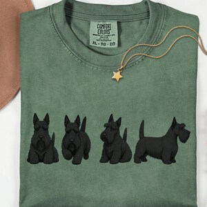 May include: Moss green t-shirt with a crew neck and short sleeves. The shirt features a graphic of four black Scottish Terrier dogs in different poses. A gold-colored necklace with a star pendant is draped on the shirt. The shirt has a Comfort Colors tag.