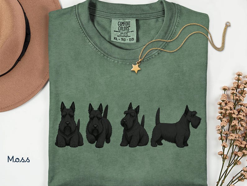 May include: Moss green t-shirt with a crew neck and short sleeves. The shirt features a graphic of four black Scottish Terrier dogs in different poses. A gold-colored necklace with a star pendant is draped on the shirt. The shirt has a Comfort Colors tag.
