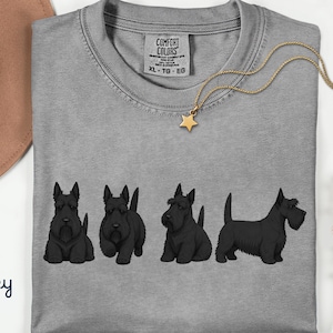 May include: A grey Comfort Colors t-shirt featuring four black Scottish Terrier dog illustrations. A gold necklace with a star pendant is draped across the shirt. The word "Grey" is written in cursive in the bottom left corner.