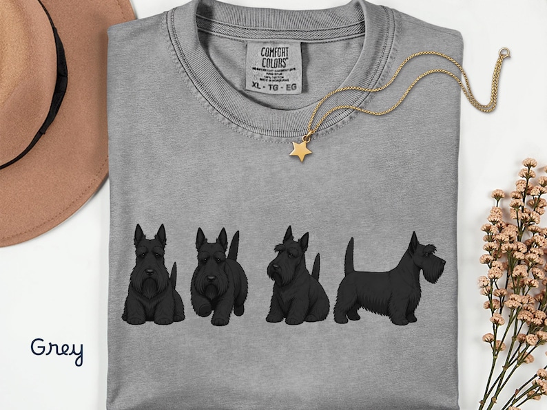 May include: A grey Comfort Colors t-shirt featuring four black Scottish Terrier dog illustrations. A gold necklace with a star pendant is draped across the shirt. The word "Grey" is written in cursive in the bottom left corner.