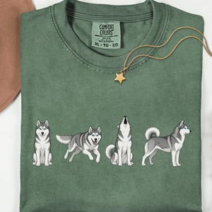 May include: Moss green t-shirt featuring a line of four cartoon huskies in different poses. The shirt has a crew neck and short sleeves. A gold necklace with a star pendant is draped on the shirt. The text "Comfort Colors" is visible on the tag.