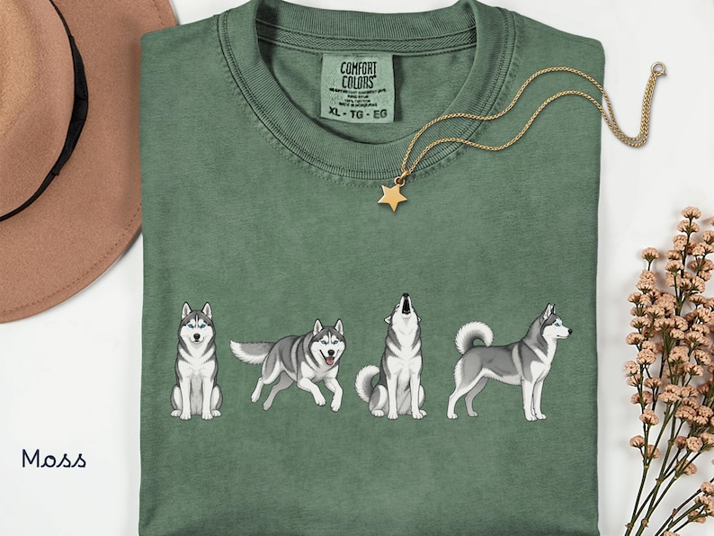 May include: Moss green t-shirt featuring a line of four cartoon huskies in different poses. The shirt has a crew neck and short sleeves. A gold necklace with a star pendant is draped on the shirt. The text "Comfort Colors" is visible on the tag.