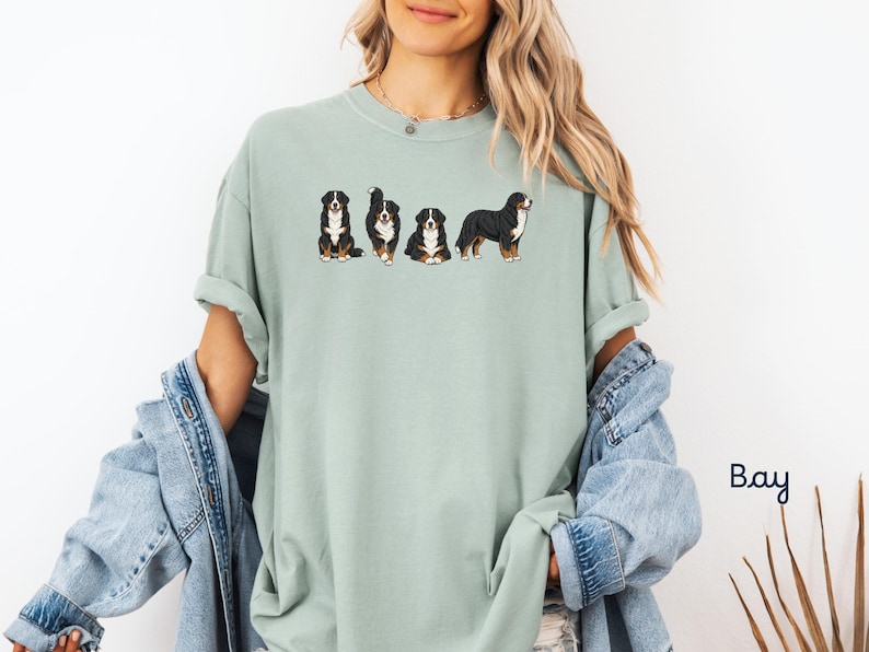 Op de afbeelding: Lichtgroen T-shirt met vier illustraties van Berner Sennenhonden in verschillende poses. De honden zijn zwart, wit en bruin. Het shirt wordt gedragen door een persoon met blond haar en een spijkerjasje.