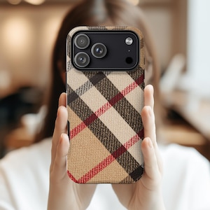 Puede incluir: Una funda de teléfono con un patrón de cuadros beige, marrón y rojo. La funda tiene un módulo de cámara negro con tres lentes y un flash. La funda se sostiene frente a una persona.