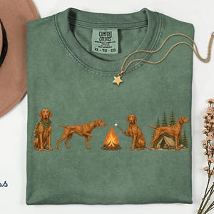 Könnte beinhalten: Ein moosgrünes T-Shirt mit einem Grafikdruck von vier Vizsla-Hunden in einer Camping-Umgebung. Die Hunde sind abgebildet, wie sie mit einem Rucksack sitzen, laufen, ein Marshmallow über einem Lagerfeuer rösten und neben einem Zelt stehen.