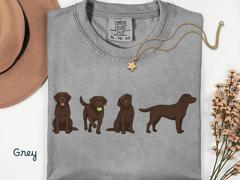 K&ouml;nnte beinhalten: Ein graues T-Shirt mit einem Grafikdruck von vier braunen Labrador-Retrievern in verschiedenen Posen, darunter einer mit einem gelben Tennisball. Das Shirt hat ein Comfort Colors Markenetikett am Halsausschnitt und ist in Gr&ouml;&szlig;e XL, TG, EG.