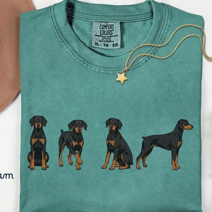 Puede incluir: Camiseta verde mar con cuatro ilustraciones de Doberman Pinschers en diferentes poses. La camiseta tiene una etiqueta Comfort Colors y un collar con colgante de estrella dorada. También se ven un sombrero marrón y flores.