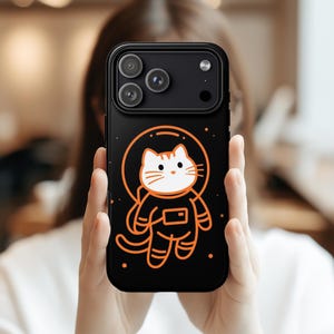 May include: Black phone case featuring a cartoon astronaut cat in an orange spacesuit. The cat is inside a circular helmet, with orange stars around it. The case has a black border and a camera module.