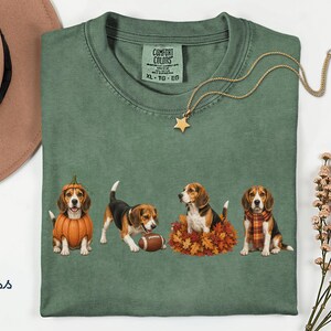 Może przedstawiać: Koszulka w kolorze mchu z grafiką przedstawiającą cztery psy rasy beagle w jesiennych strojach. Jeden beagle ma na sobie kostium dyni, drugi bawi się piłką do futbolu, trzeci siedzi w jesiennych liściach, a czwarty ma na sobie szalik w kratę.