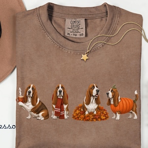 以下が含まれることがあります： 4匹のバセットハウンドが描かれた茶色のTシャツ。1匹は湯気の立つマグカップを持ち、1匹は赤いスカーフを巻き、1匹は落ち葉の中に座り、1匹はカボチャのコスチュームを着ている。Comfort ColorsのXLサイズタグ付き。