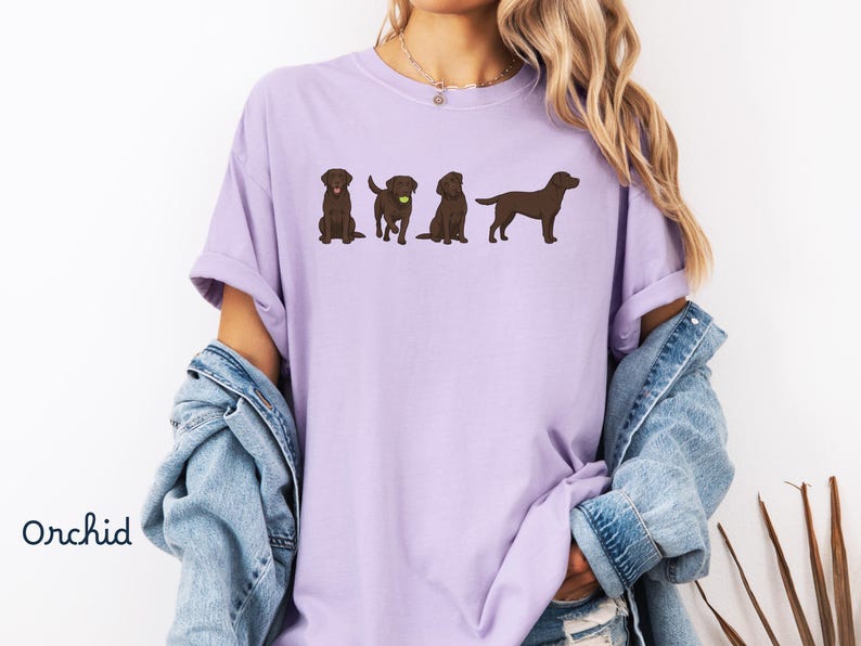 K&ouml;nnte beinhalten: Ein hellviolettes T-Shirt mit einem Grafikdruck von vier braunen Labrador-Hunden in verschiedenen Posen, darunter einer mit einem gelben Ball. Das T-Shirt hat einen Rundhalsausschnitt und kurze &Auml;rmel, mit dem Wort Orchid unten links.