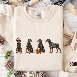 May include: A sand-colored crewneck sweatshirt featuring a graphic of four Doberman dogs. One wears a Santa hat, one is wrapped in colorful Christmas lights, one sits by a red gift box, and one wears a green sweater with white snowflake patterns.