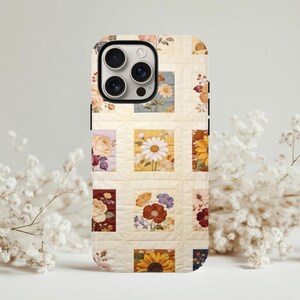 Könnte beinhalten: Eine Handyhülle mit einem Patchwork-Quilt-Design mit floralen Mustern. Die Hülle hat einen cremefarbenen Hintergrund mit quadratischen Blöcken, die verschiedene Blumen wie Gänseblümchen, Rosen und Sonnenblumen in Gelb-, Rot- und Blautönen zeigen.