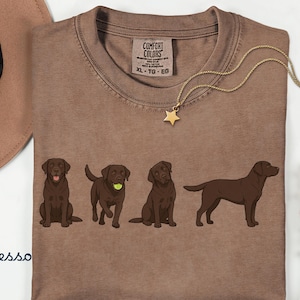 K&ouml;nnte beinhalten: Ein espressofarbenes T-Shirt mit einem Grafikdruck von vier schokoladenbraunen Labrador-Retrievern in verschiedenen Posen. Das Shirt hat einen Rundhalsausschnitt und ein Comfort Colors Markenetikett am Innenkragen mit der Gr&ouml;&szlig;enangabe XL.
