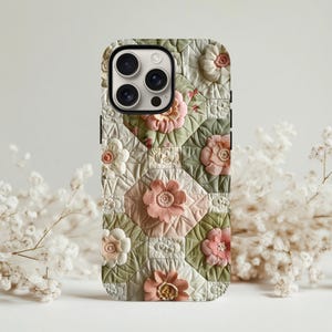 May include: A phone case featuring a quilted floral design. The case has a pattern of pink, cream, and green flowers arranged in a geometric pattern. The case has a black border and a camera cutout.
