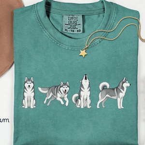 May include: Seafoam green t-shirt featuring a graphic of four Siberian Huskies in different poses. The shirt has a crew neck and short sleeves. A gold necklace with a star pendant is draped on the shirt. The shirt has a tag that says "Comfort Colors".