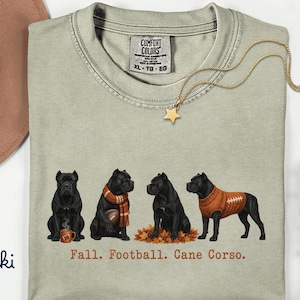 Könnte beinhalten: Ein khakifarbenes T-Shirt mit Rundhalsausschnitt und einer Grafik von vier schwarzen Cane Corso Hunden. Ein Hund sitzt mit einer Tasse, einer trägt einen Schal mit Football, einer sitzt in Herbstlaub und einer trägt ein Trikot. Text: Fall. Football. Cane Corso.