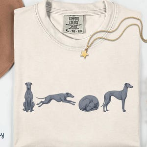 以下が含まれることがあります： アイボリー色のTシャツ。グレーハウンドが様々なポーズで描かれたグラフィックが特徴です。「Comfort Colors」と書かれたタグが付いています。星のペンダントが付いたゴールドのネックレスがTシャツの上にかけられています。「Ivory」の文字が左下にプリントされています。