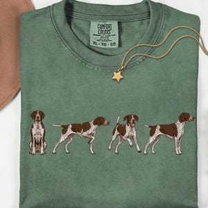 May include: Moss green t-shirt featuring a graphic of four brown and white dogs in different poses. The shirt has a crew neck and short sleeves. A gold necklace with a star pendant is draped on the shirt. The shirt is labeled "Comfort Colors".