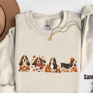 Può includere: Una felpa girocollo color sabbia con un ricamo raffigurante quattro cani Basset Hound con abbigliamento autunnale, tra cui sciarpe e berretti, circondati da zucche, tazze di caffè e foglie cadute, con una collana a forma di stella.