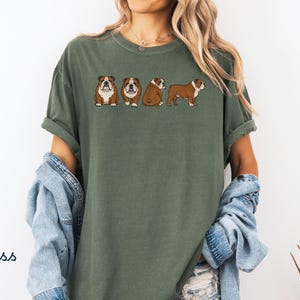 May include: A moss green t-shirt with a design of four cartoon bulldogs in different poses. The bulldogs are brown and white with black outlines. The shirt has short sleeves and a relaxed fit. The word "Moss" is printed in the bottom left corner.